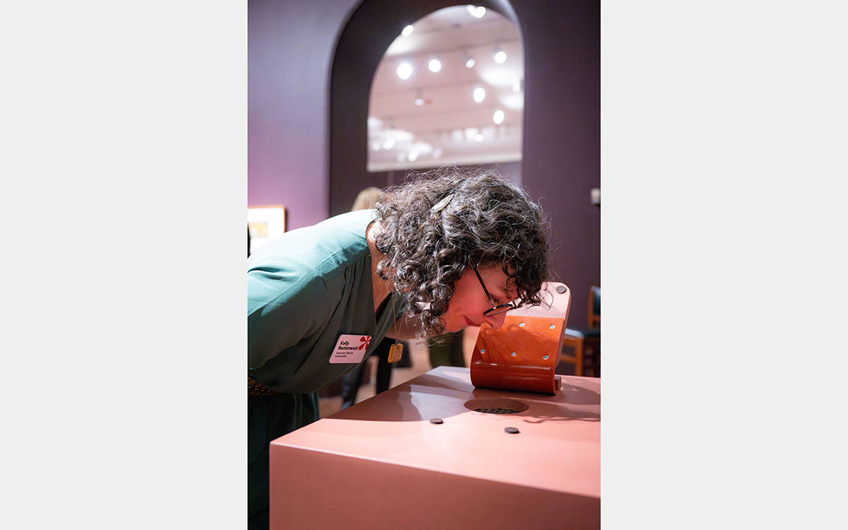 A woman lowers her head towards a table-top olfactory station; in the background is an archway leading into another room.