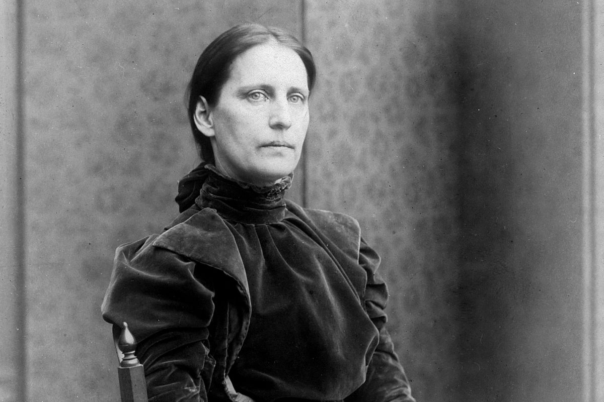 In a black and white photograph, a woman dressed in Victorian clothes stares at the viewer with a solemn expression.
