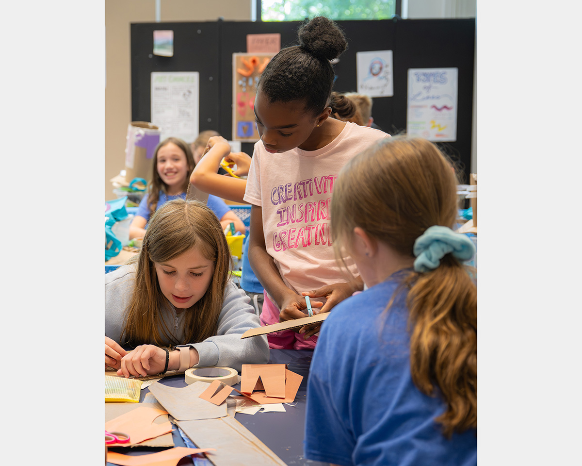 Three girls gather around a table making art while a fourth girl smiles in the background.