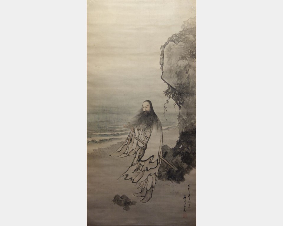 A long-bearded man, dressed in boots and a tattered robe, stands on a beach below a rock formation.
