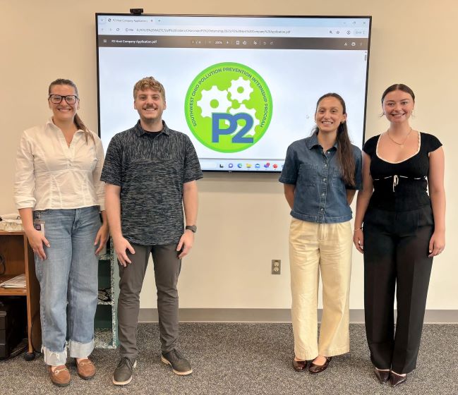 Three women and a man stand by a presentation screen.