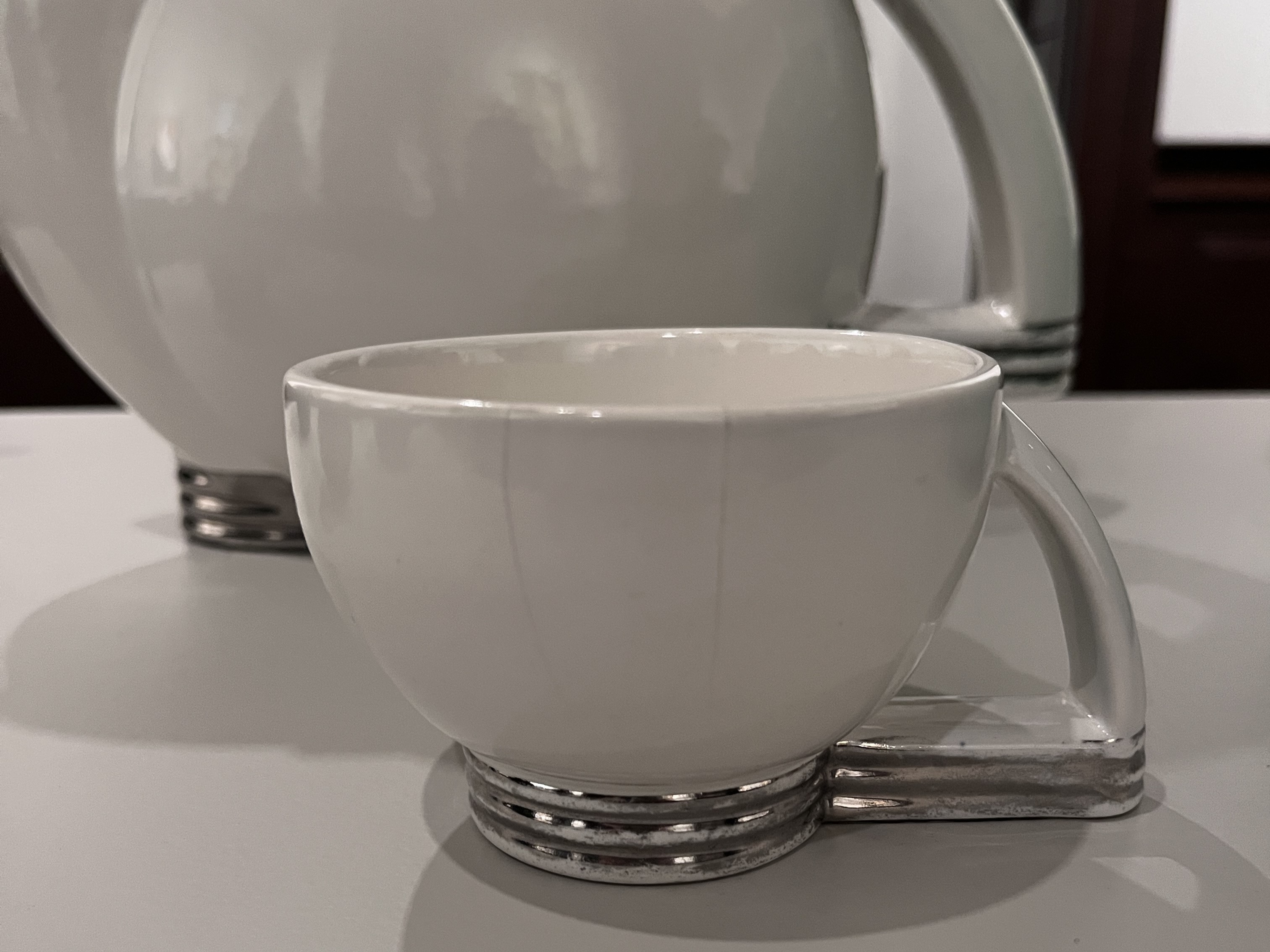 white ceramic teacup with chrome-colored ribbed base and angular handle with base of pitcher in background