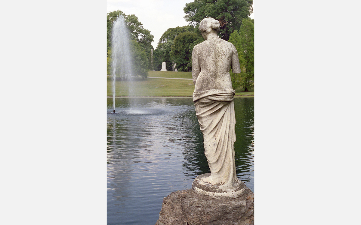 A marble sculpture of the back of a woman overlooking a lake with fountain in a green park-like setting.