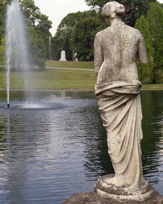 A marble sculpture of the back of a woman overlooking a lake with fountain in a green park-like setting.