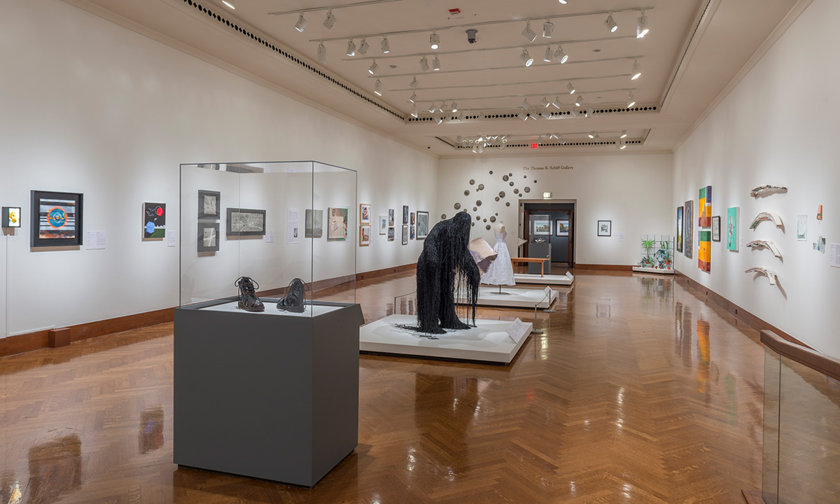 A gallery full of art including boots, a large dark figure draped in strings, and a dress
