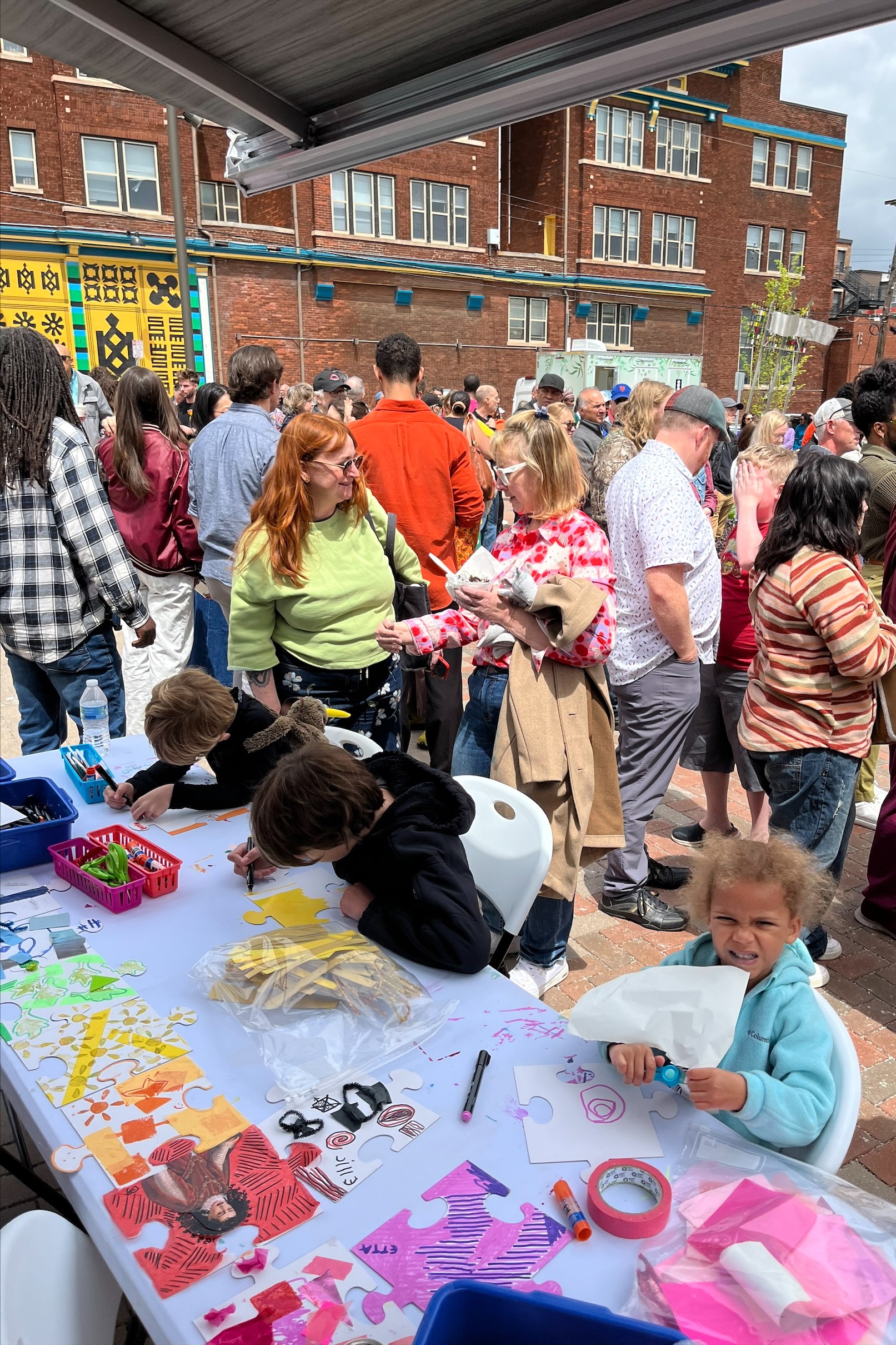 Three kids make art on a table with a crowd of people in the background and a large brick building beyond.