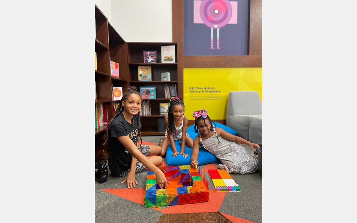 Three girls sit on the floor surrounding a colorful puzzle in a room with a bookcase and soft furniture.
