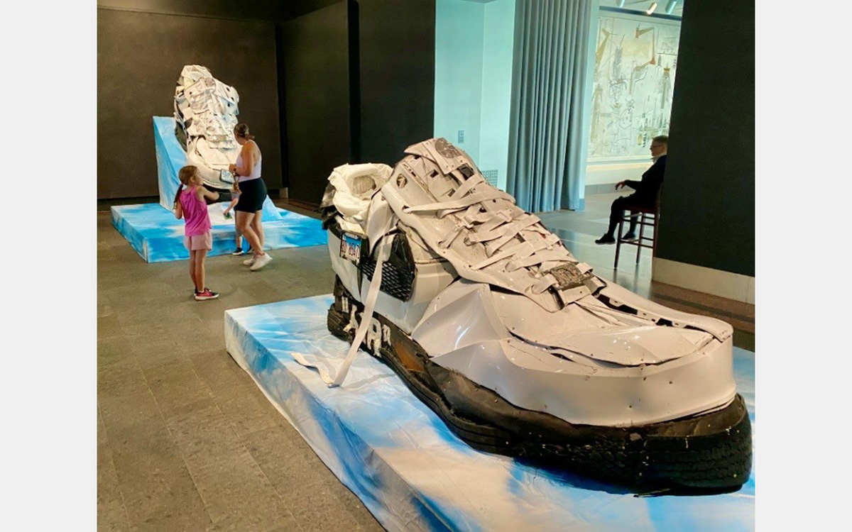 Large-scale metal sculptures representing a pair of sneakers displayed inside a gallery space with visitors and a security guard sitting in a chair