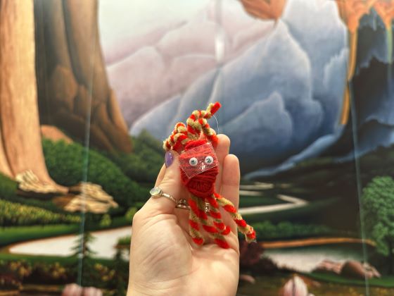 A human hand holds an animal-like figure made of yard against the backdrop of a landscape painting.