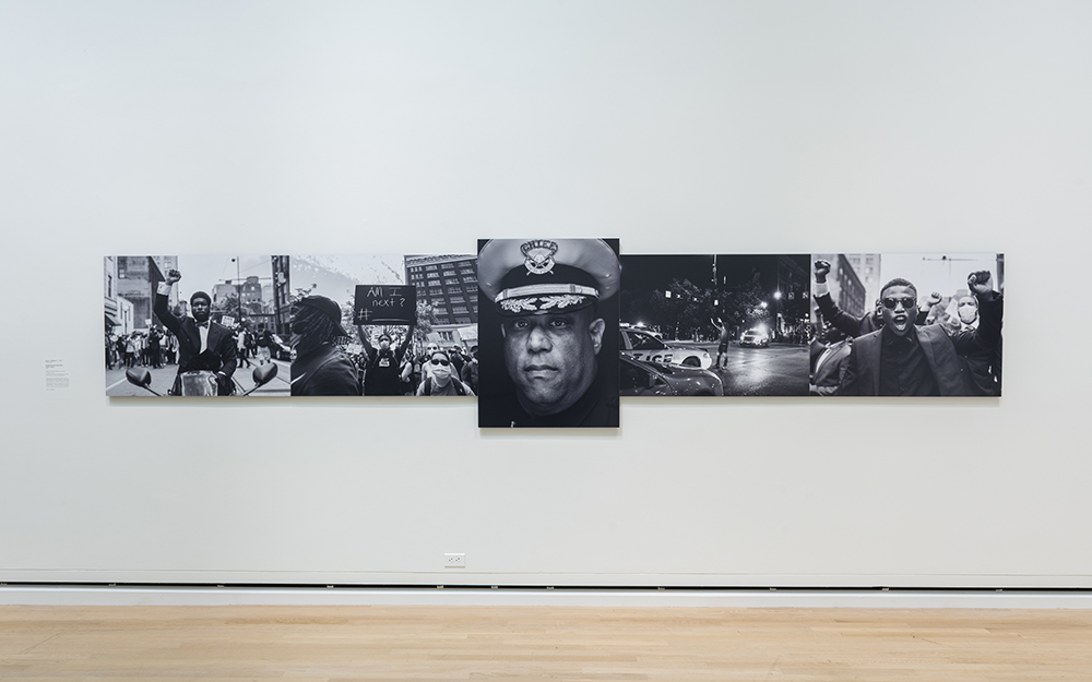Kevin J. Watkins' Arrest the cops that killed (insert name), a series of black and white photographs of African Americans in protest. The center photograph is a close up portrait of an African American police chief in uniform