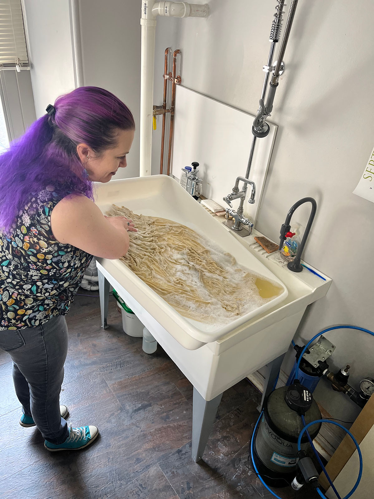 A white woman with purple hair washes the yellowed blouse and skirt in a large utility sink