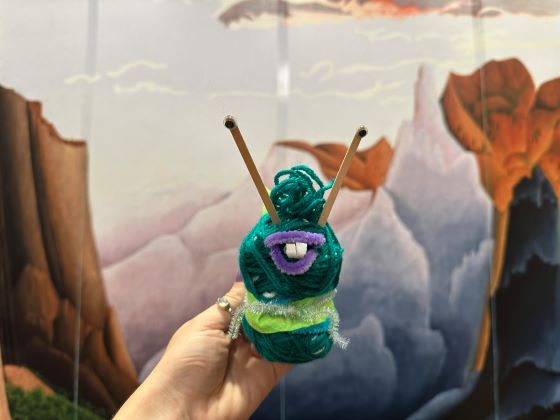A human hand holds an animal-like figure made of yard against the backdrop of a landscape painting.