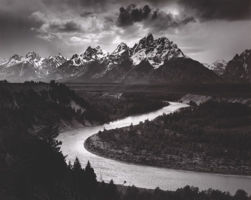 A black and white photo of a twisting river in front of a jagged mountain range