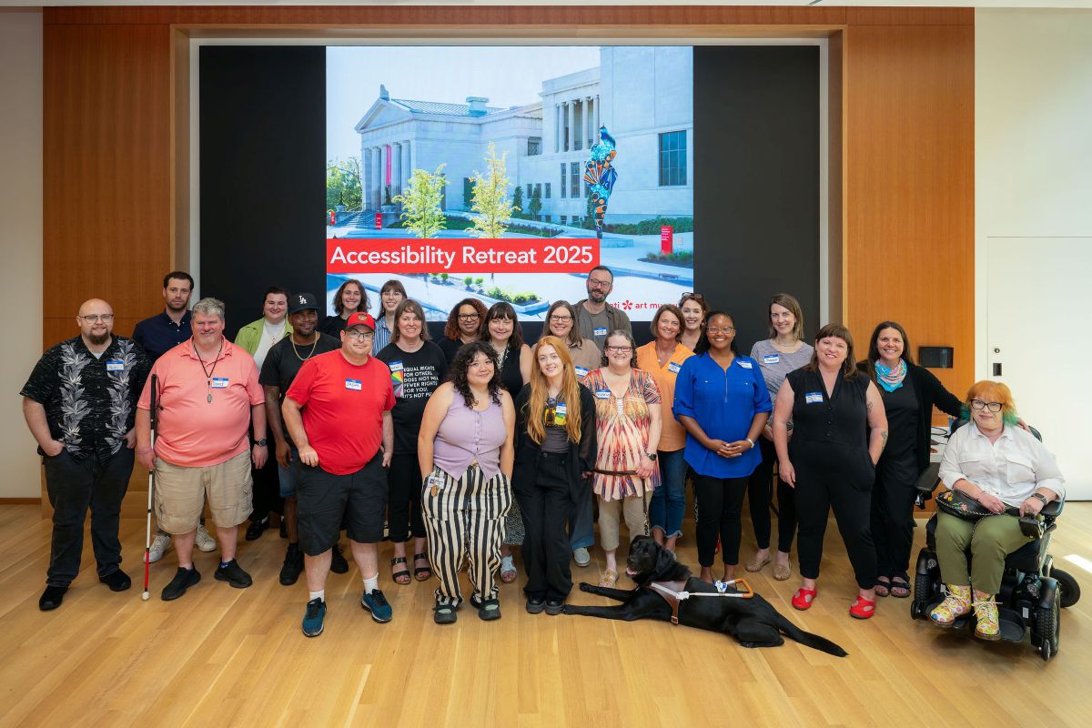 A group of 23 people (one sitting in a wheelchair) and a service dog pose in front of a large project screen stating "Accessibility Retreat 2025"