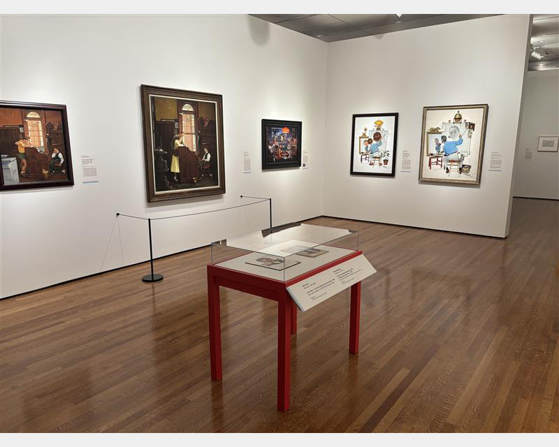 Interior view of a gallery with white walls and hardwood floors. Five framed artworks hang on the walls and table-top display case with red legs stands in the center of the room.
