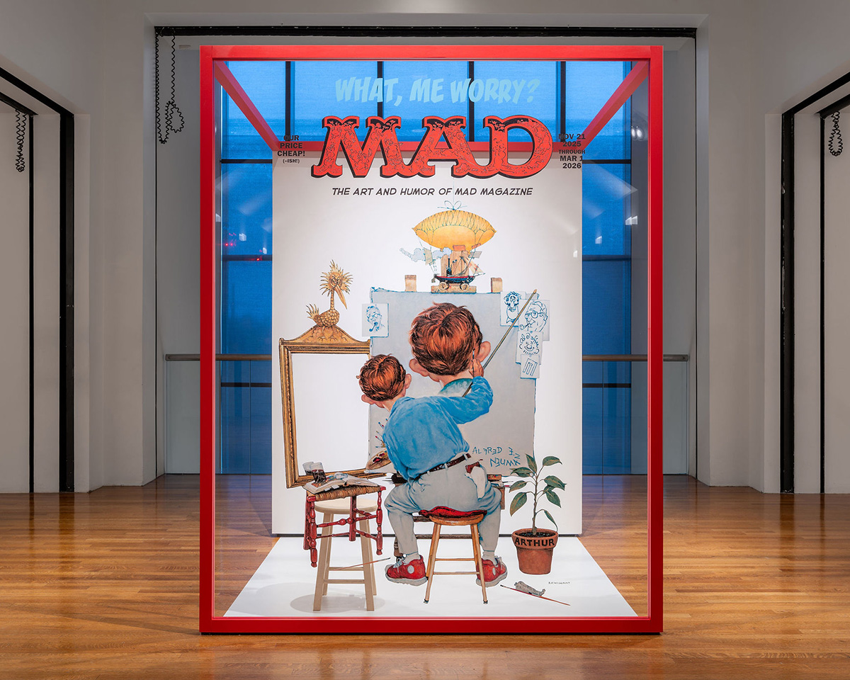  A large transparent sign, framed in bright red, features an illustration of a guy sitting on a stool and painting at an easel.  The sign is back-lit and stands on a wood floor in a room with white walls.