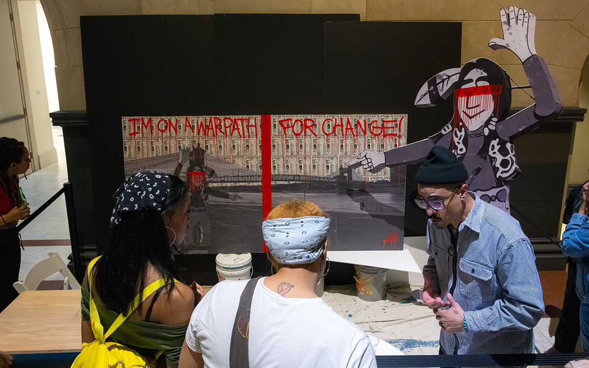 An indigenous man talks to two visitors about an artwork that reads "I'm on a warpath for change!"