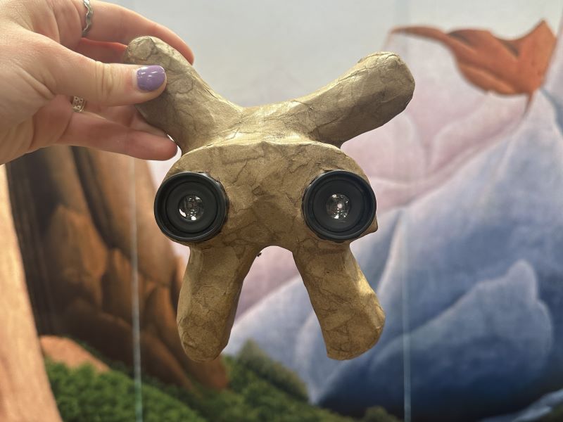 Hand holding binoculars encased in paper maché with landscape painting in background.