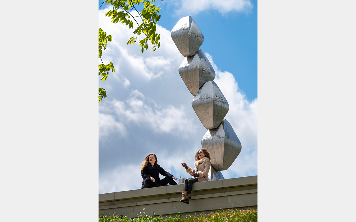 Two women sitting on a stone wall beneath a contemporary steel sculpture.