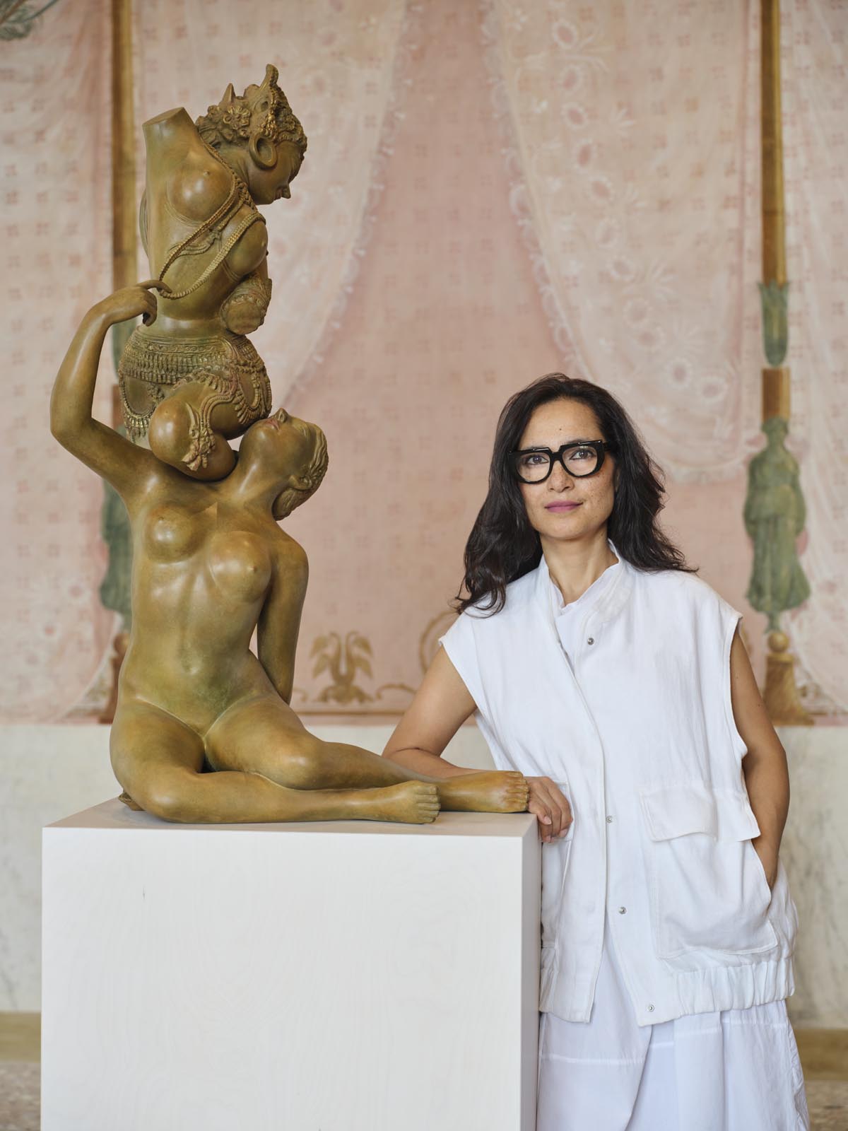 Shahzia Sikander wears a white outfit and dark rimmed glasses while leaning on a plinth featuring a brown sculpture of two nude women