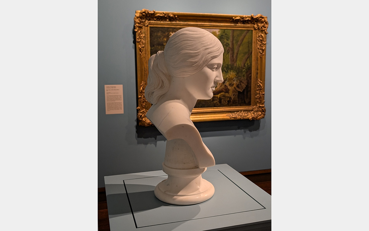 A white marble bust of a woman with a painting in gold frame in the background.
