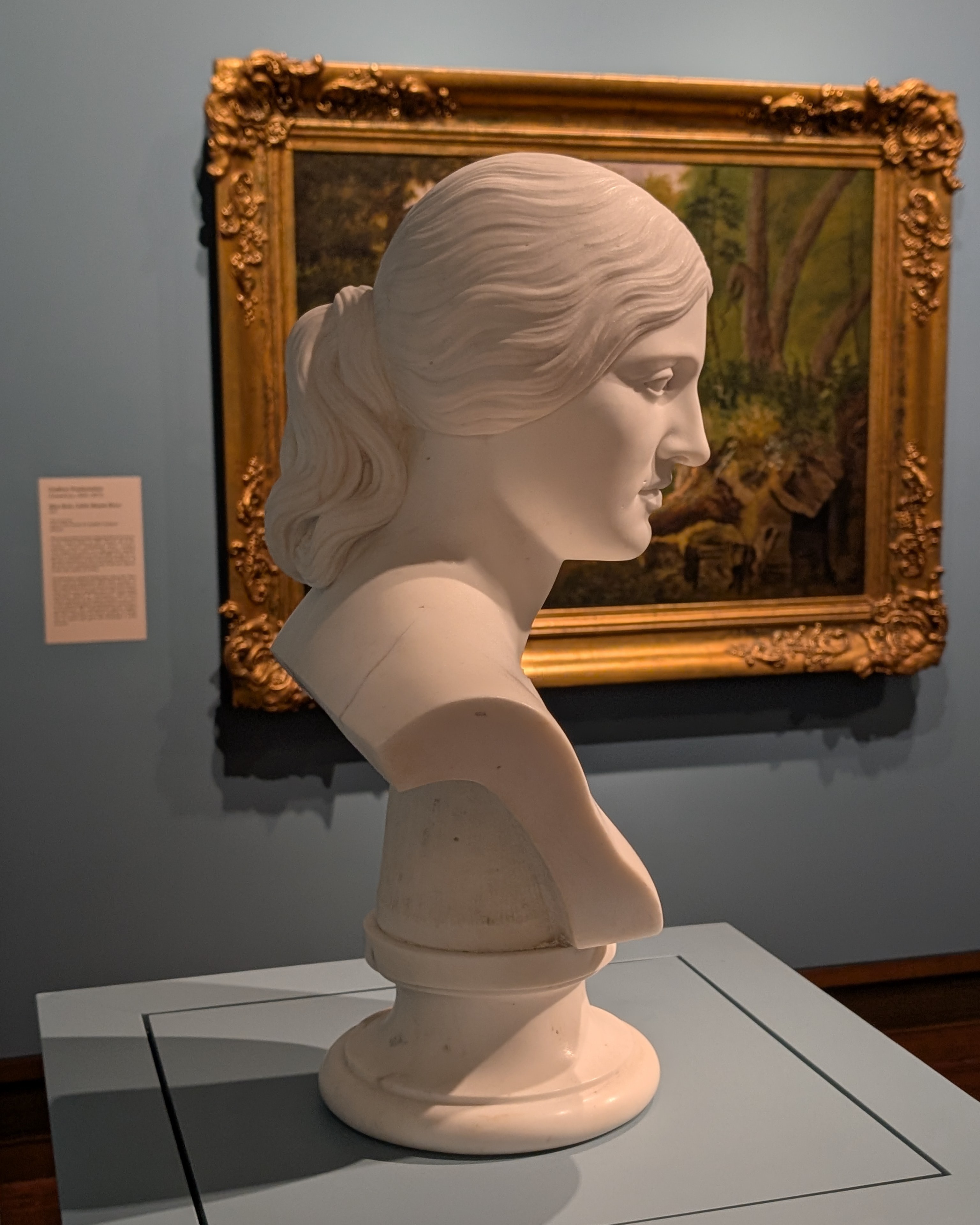 A white marble bust of a woman with a painting in gold frame in the background.