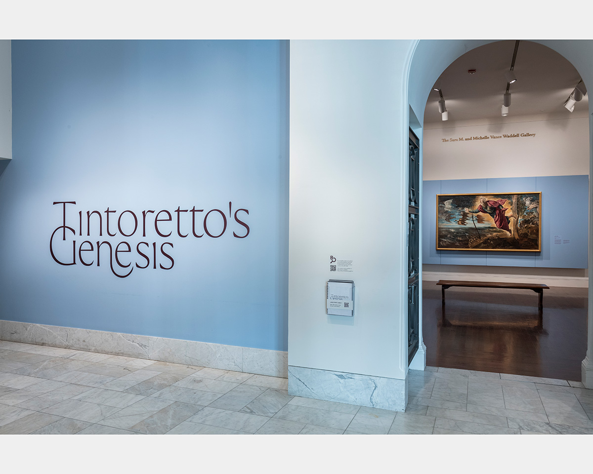 An archway punctures a blue wall with lettering stating, "Tintoretto's Genesis." Through the archway, inside a gallery space, a colorful painting hangs on the back wall in front of a blue wall.