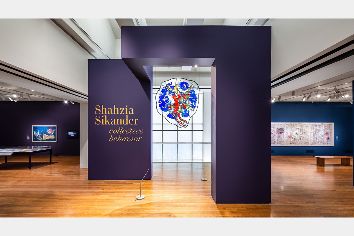 A large red and blue glass piece hangs from a purple wall that says "Shahzia Sikander: Collective Behavior"