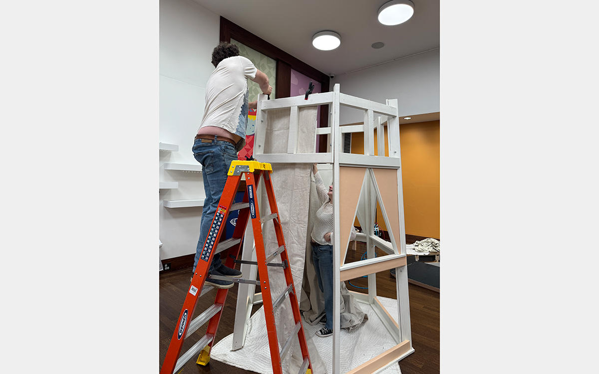 Two people, one on an orange ladder, building a wood structure inside a room.