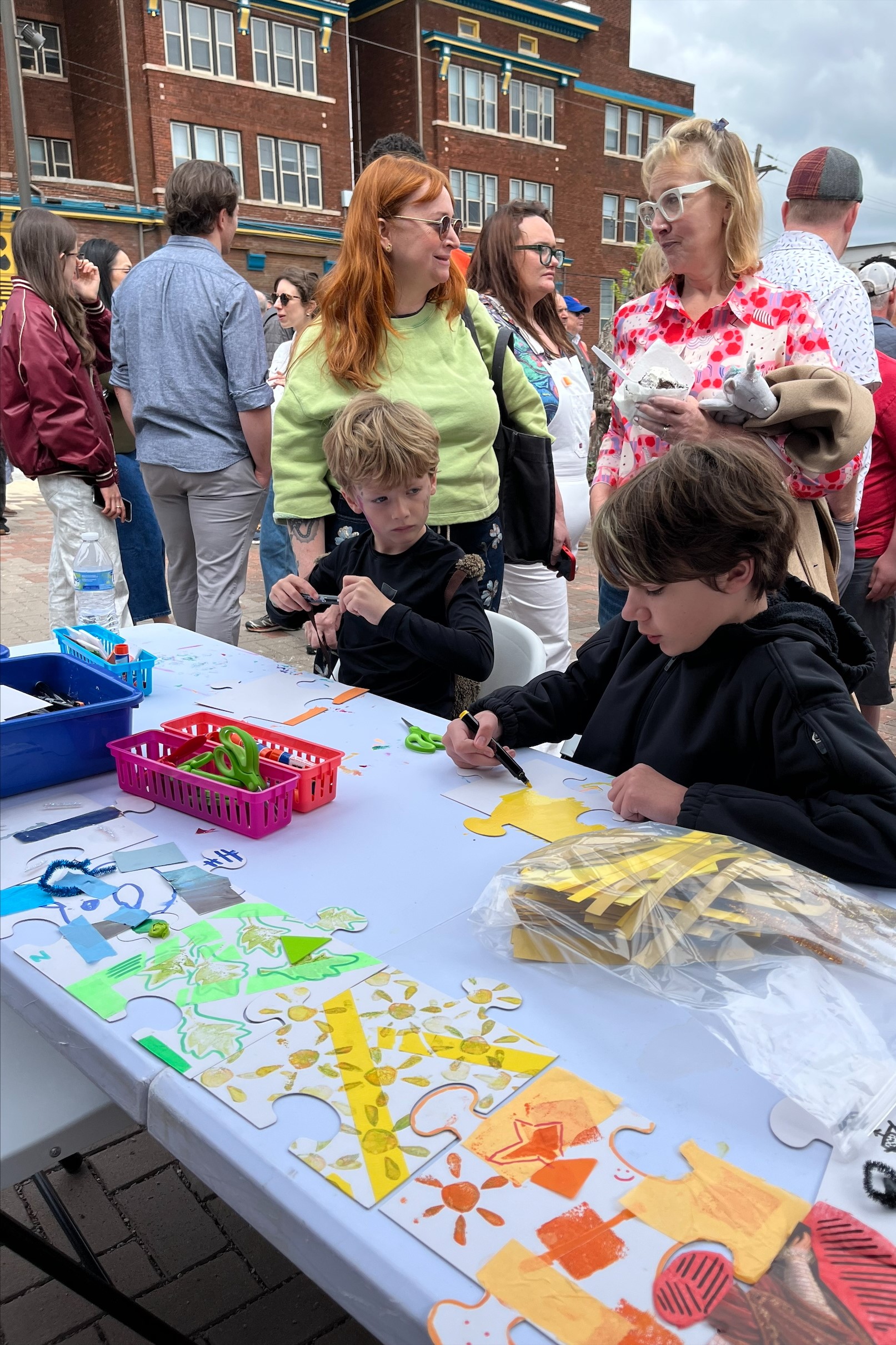 Two boys make art at a table with a crowd of people in the background and a red brick building beyond.