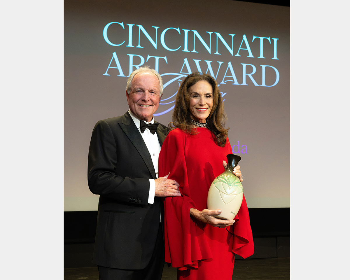 Two people, one dressed in a black tuxedo and the other dressed in a bright red gown, stand in front of a projection screen stating, "CINCINNATI ART AWARD". The woman in the red dress hold a vase.