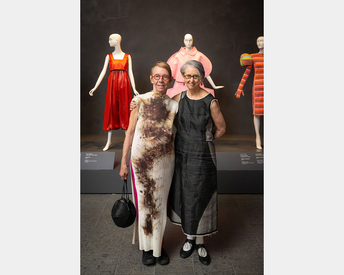 Two women in long dress stand in front of a platform with mannequins wearing bright-colored dresses.
