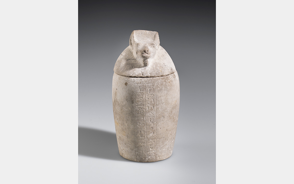 Buff colored stone jar with a jackal-like head for the lid and hieroglyphic writing down the center of the jar.
