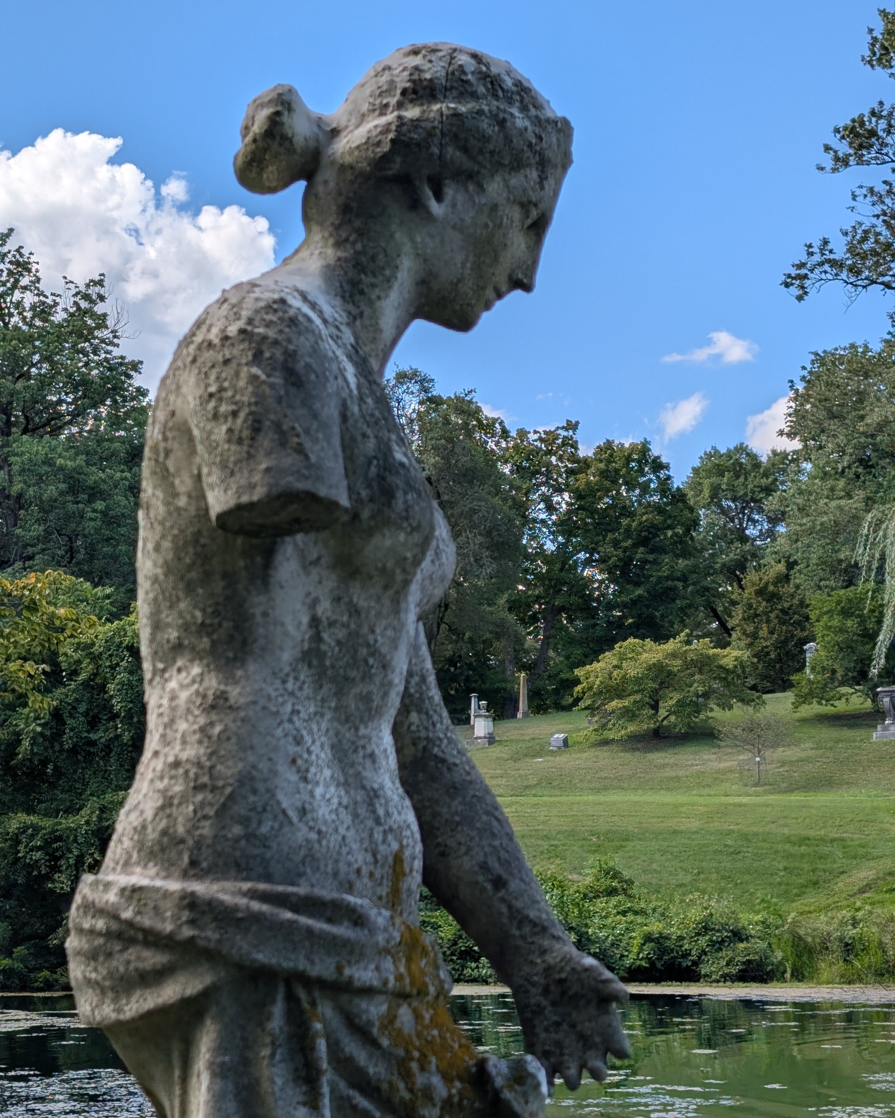 A marble sculpture of a woman wearing drapery from the waist down and nude from the waste up. A green landscape with trees, pond and ,blue sky is in the background.
