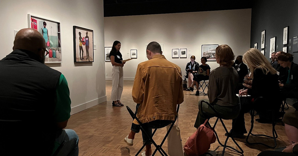 A group of visitors sit in a semicircle around a presenter, who stands by two framed photographs hanging on a wall.