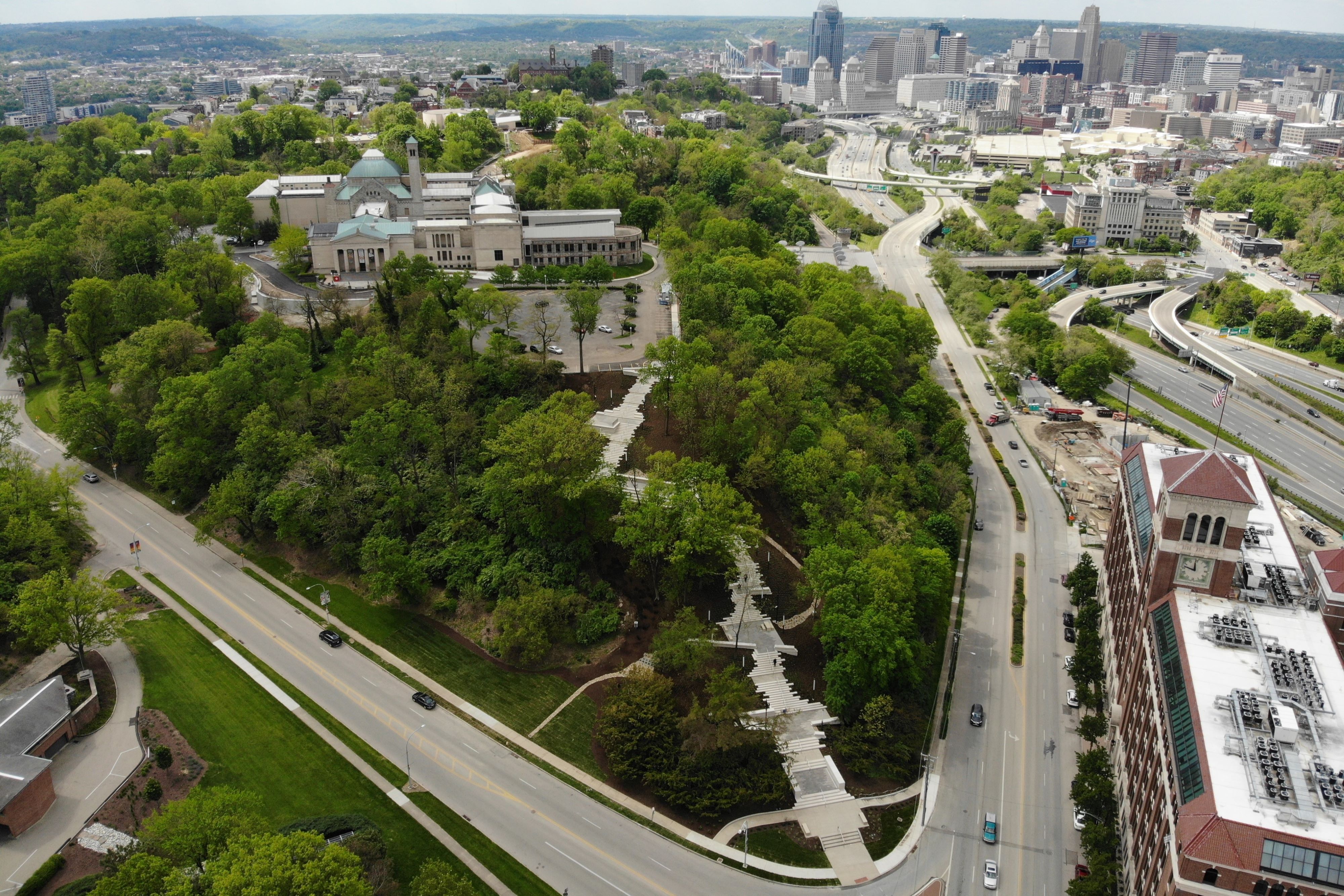 A series of steps and landings climb a wooded hillside connecting a roadway intersection with a museum on a hilltop. A city skyline is visible in the background.
